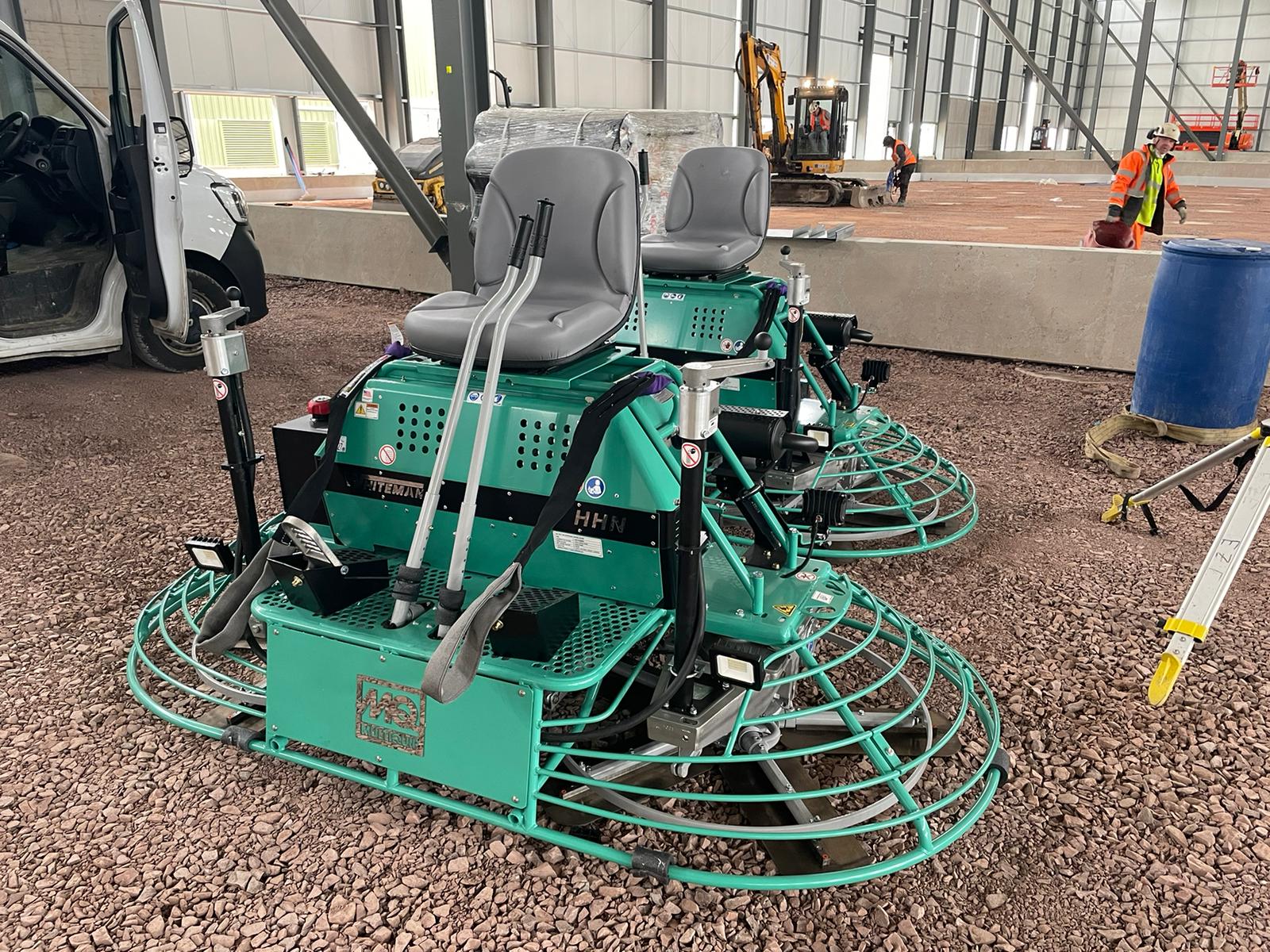 Two green power trowels for concrete finishing stored indoors at a construction site, with workers in the background.